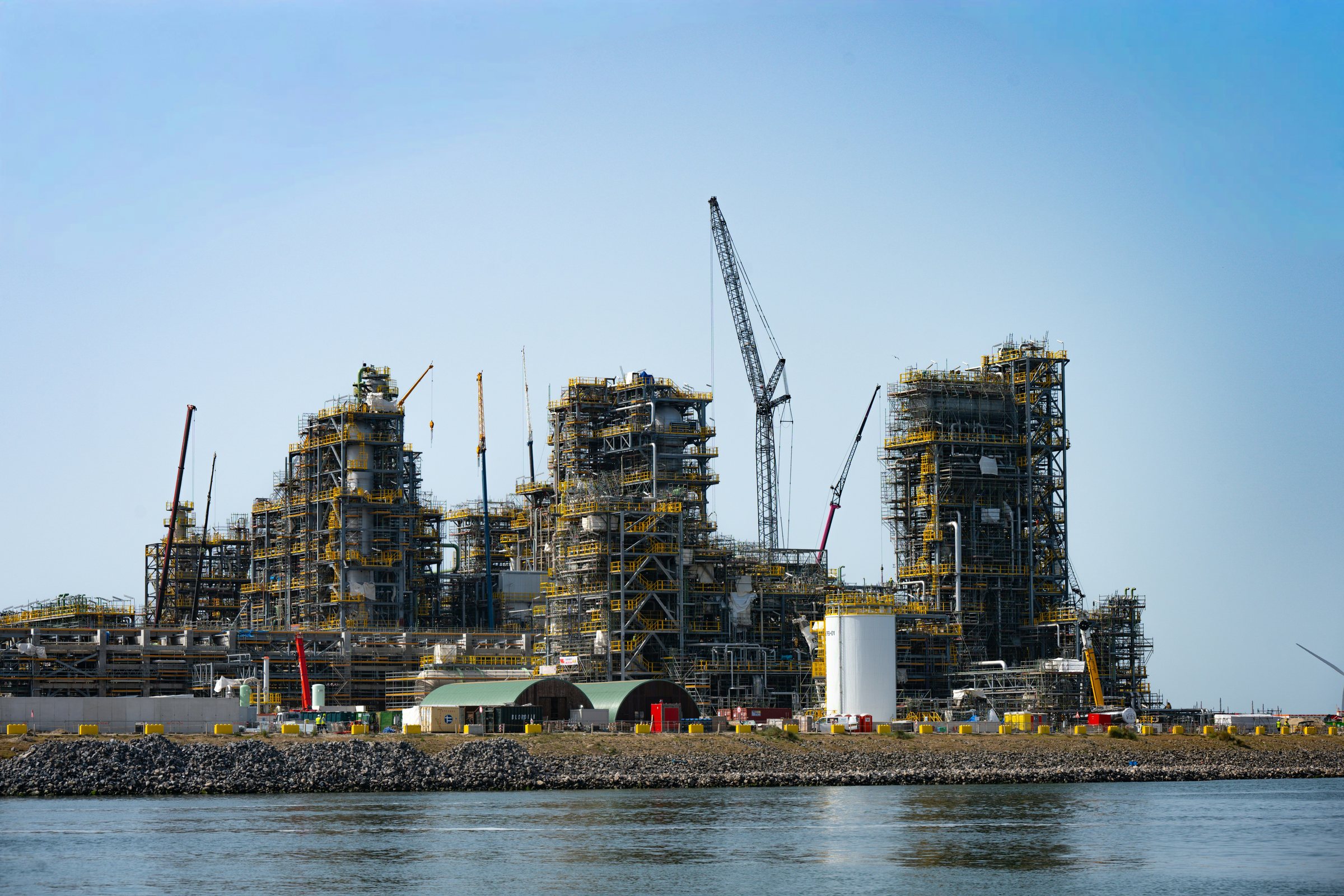 Large process plant under construction on a waterfront, with structural modules and cranes
